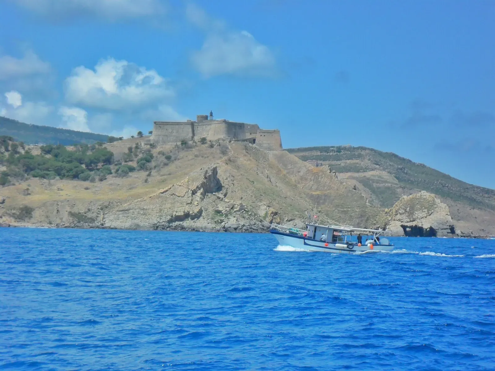 A small boat passing below Tabarka's headland fort, blue sea in the foreground — Cap Tabarka.