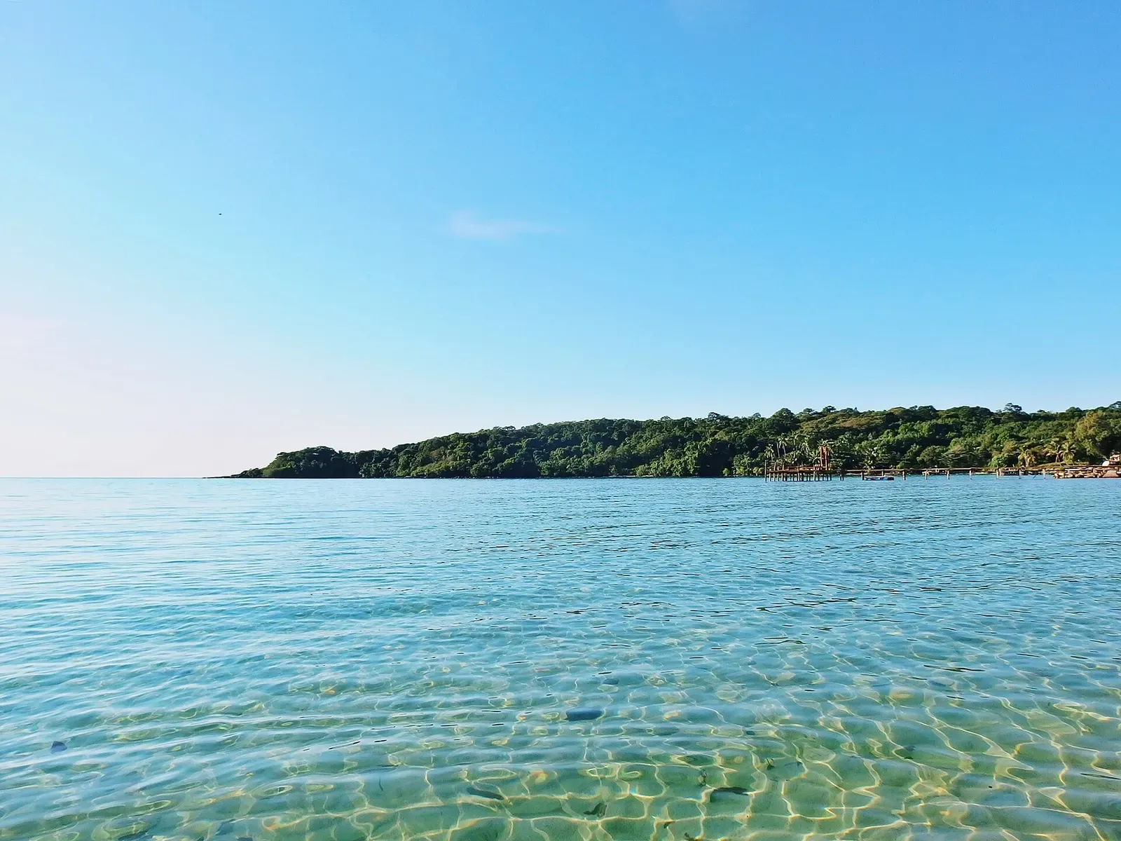Clear, shallow turquoise water over a sandy bottom, with a low forested headland on the horizon under a blue sky — Koh Kood & Koh Mak.