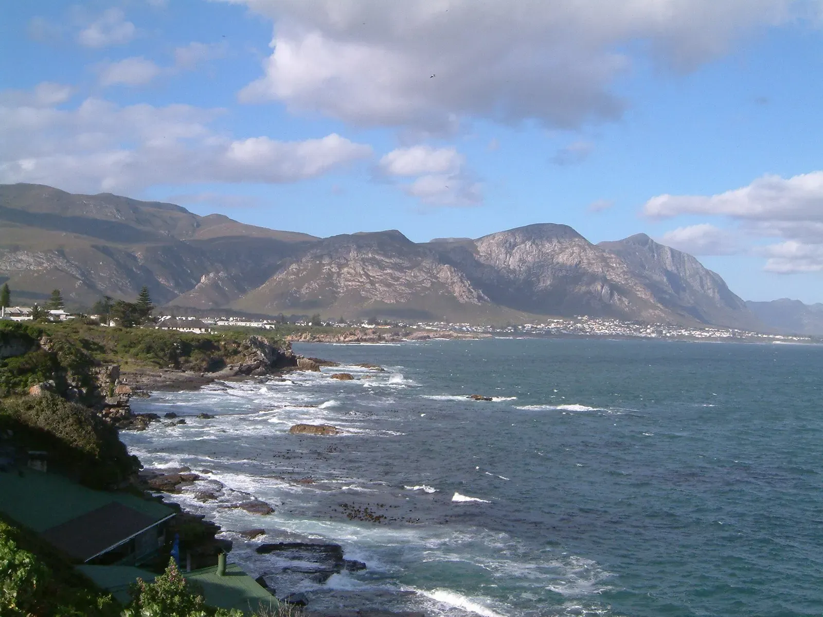 Rocky shoreline with waves breaking below a town at the foot of rugged mountains, under a blue sky with white clouds — Hermanus.