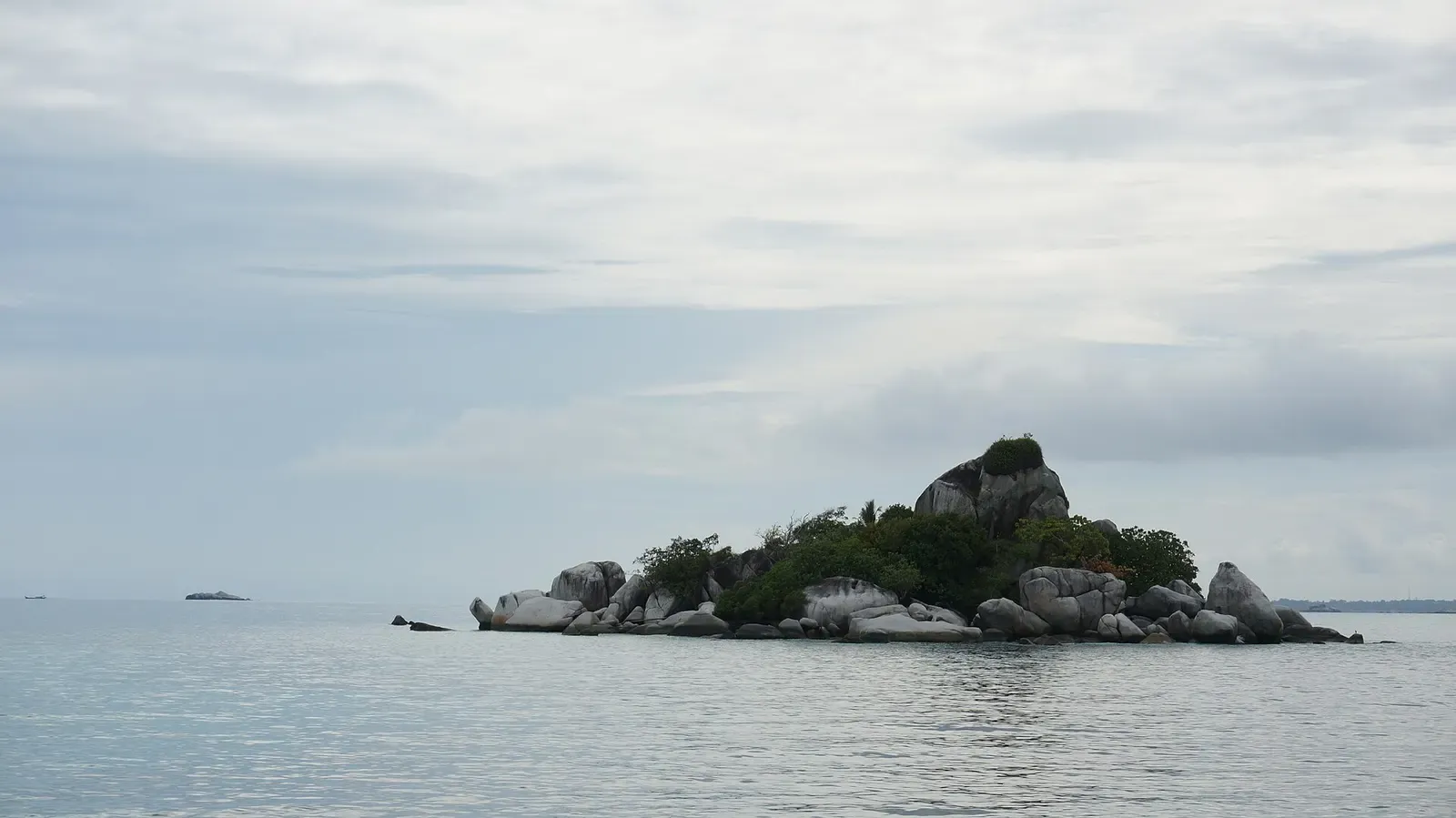 A small wooded islet ringed with grey granite boulders in a calm, pale sea under an overcast sky — Belitung