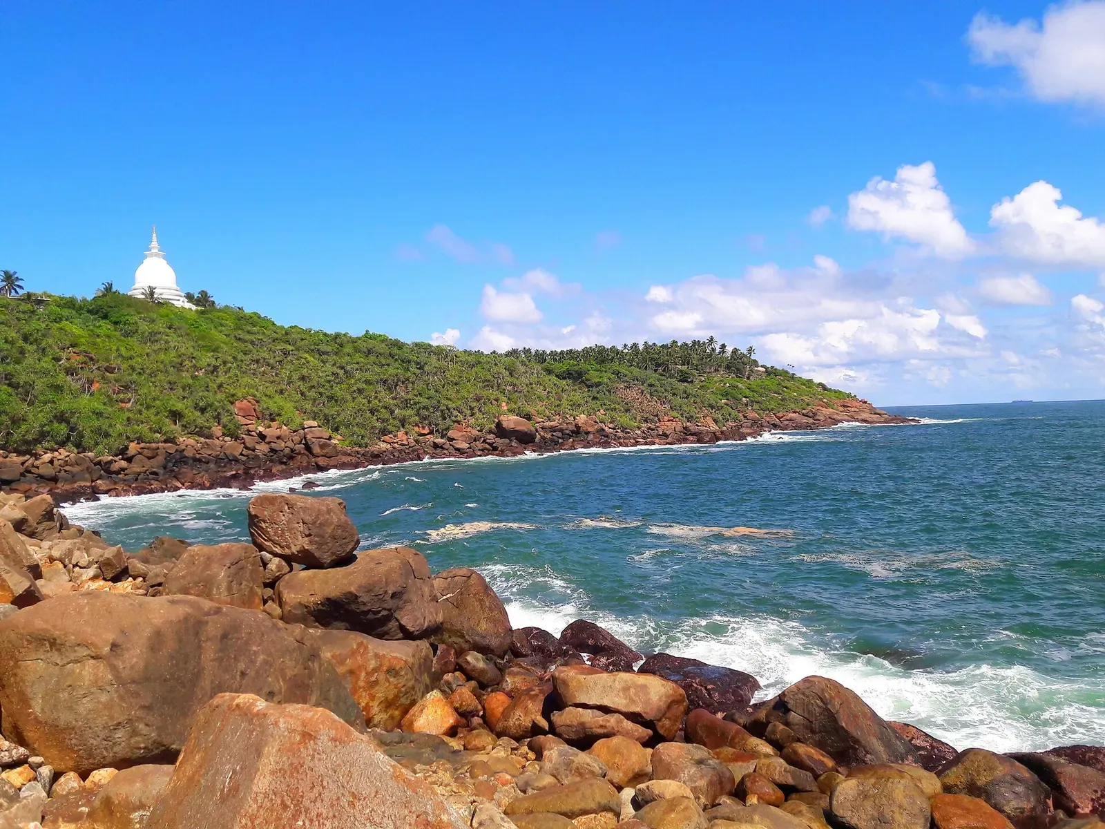 Rocky shore at Jungle Beach below a green headland topped by a white stupa, with waves breaking on red-brown boulders