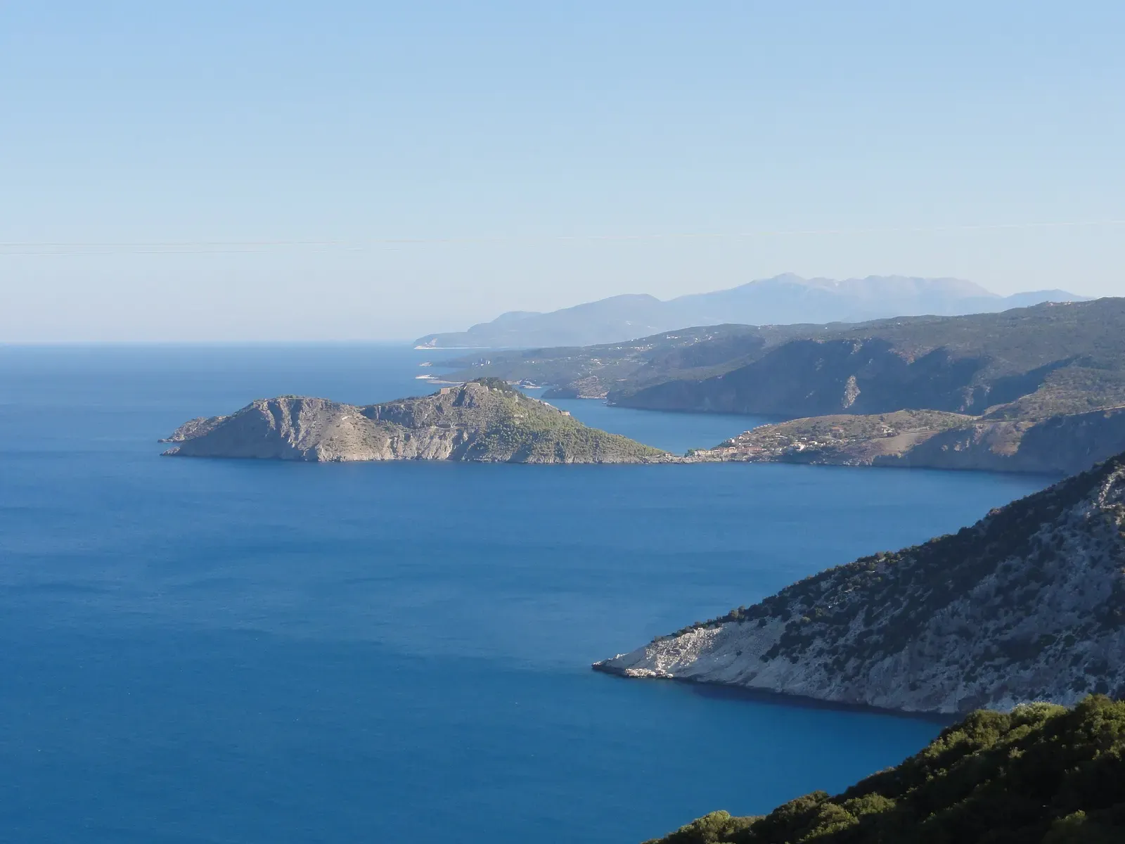 Steep Kefalonia coastline dropping into deep blue water, with a rocky headland and hazy mountains behind