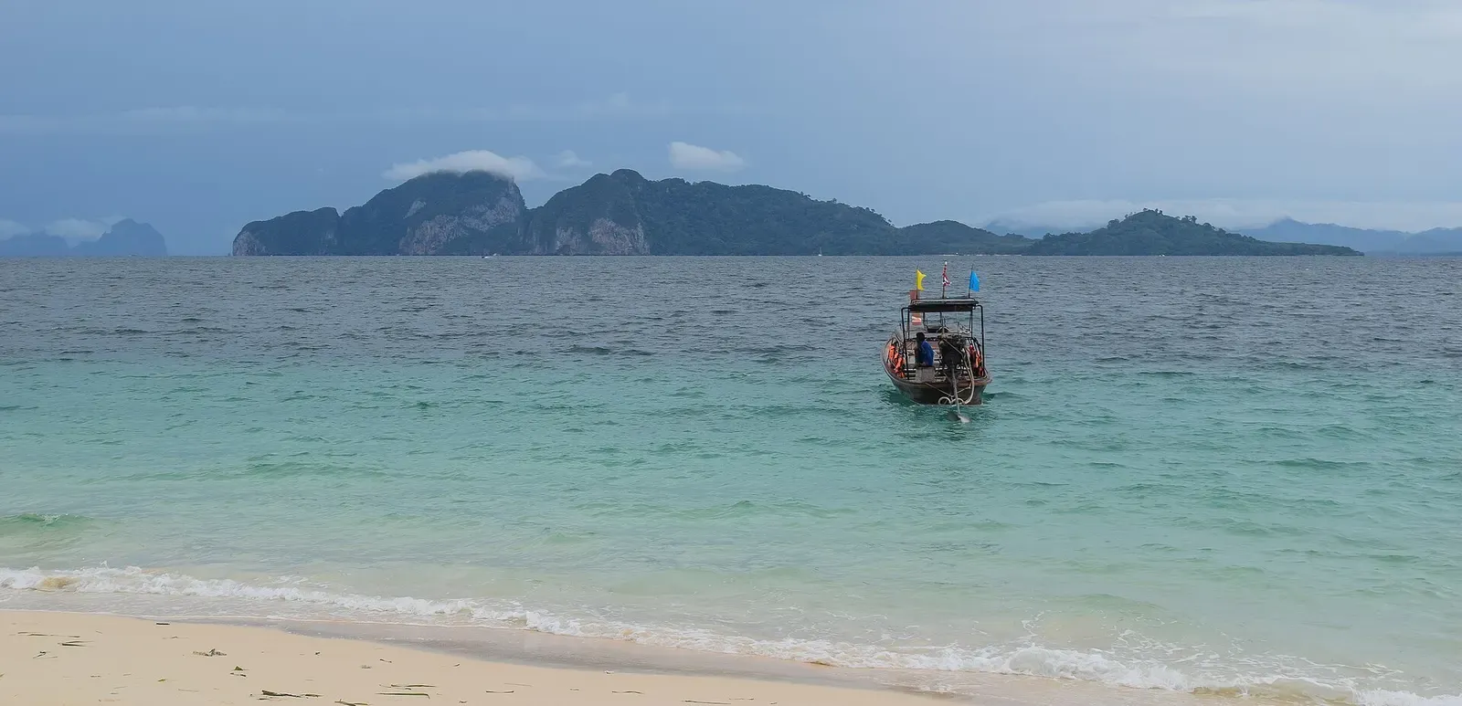A wooden longtail boat moored in turquoise shallows off a sandy beach, with a long rocky island on the horizon — Trang Islands