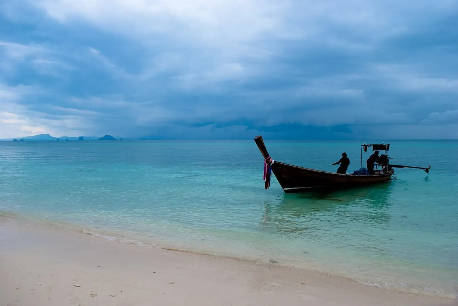 Traditional longtail boat anchored off a white-sand beach under a stormy sky