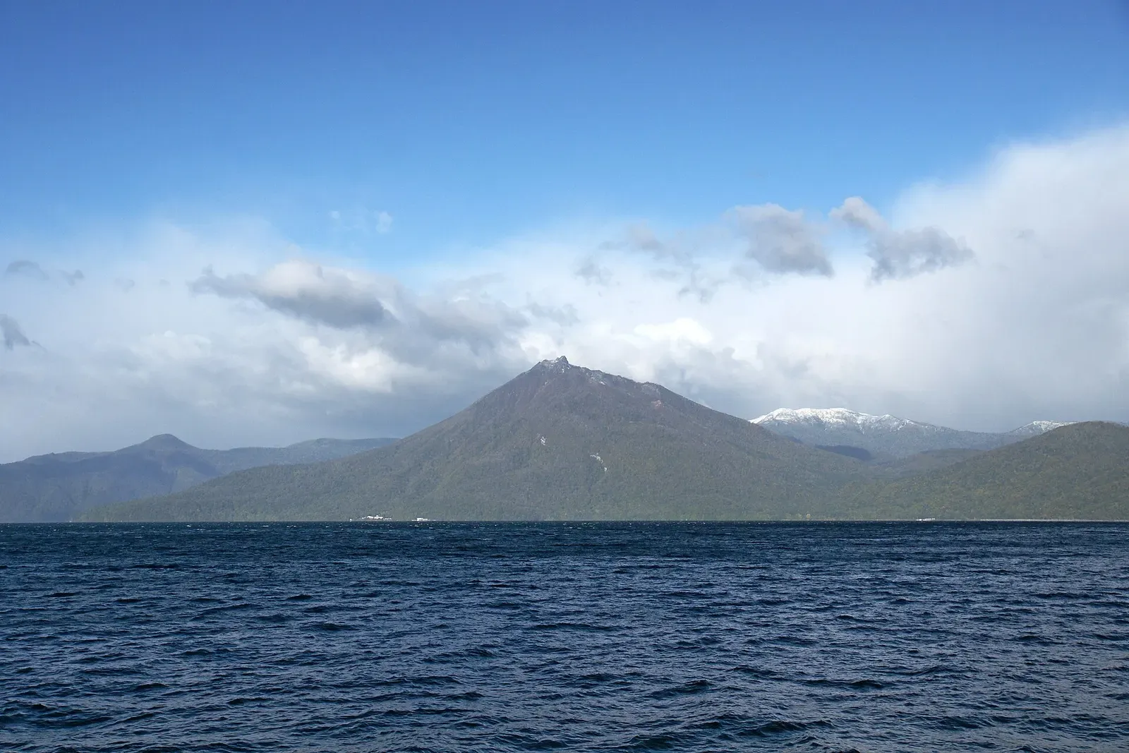 A forested cone-shaped mountain rises behind dark blue choppy water under a blue sky with white cloud — Lake Shikotsu