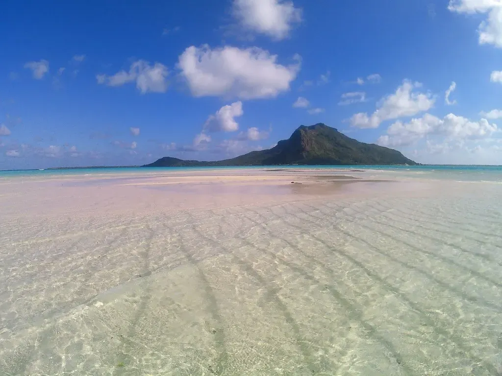 Clear, ankle-deep water over rippled white sand in the foreground, with a single green peak rising across the lagoon under a blue sky — Maupiti.