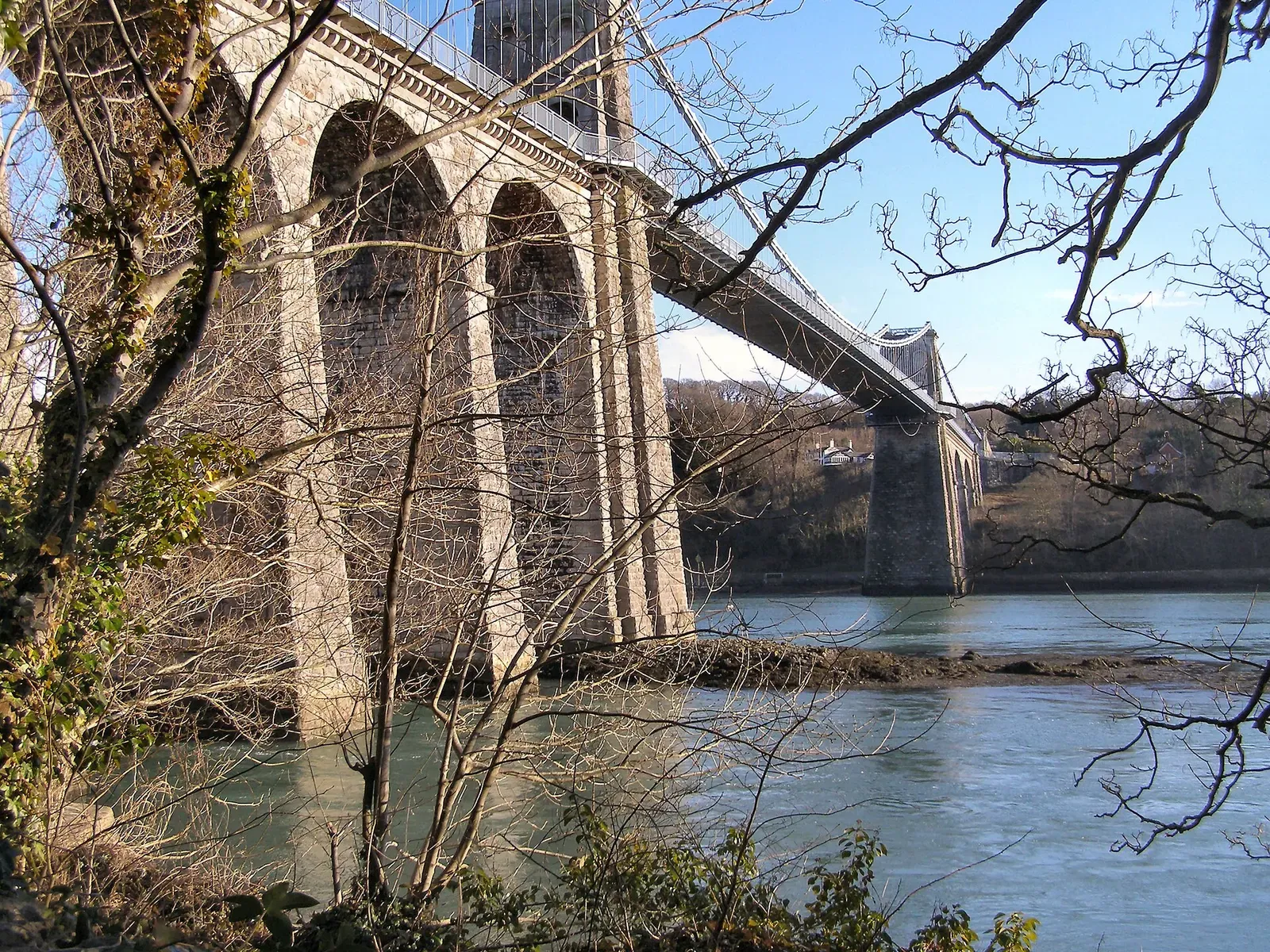 Massive stone arches of a suspension bridge rising from the water beside the shore — Menai Suspension Bridge, Anglesey