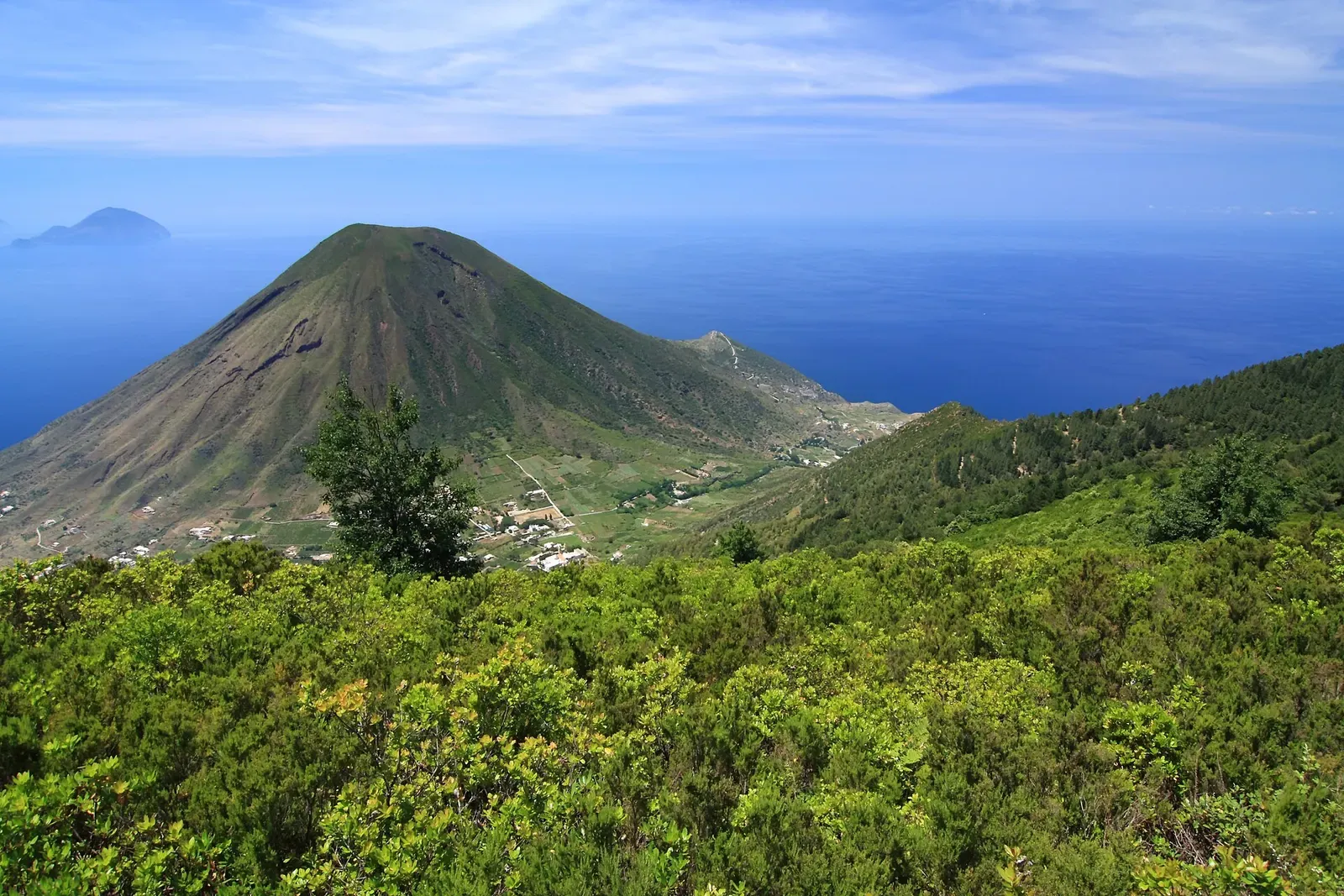 A green cone-shaped volcano above dense scrub, with blue sea and a distant island on the horizon — Filicudi & Salina