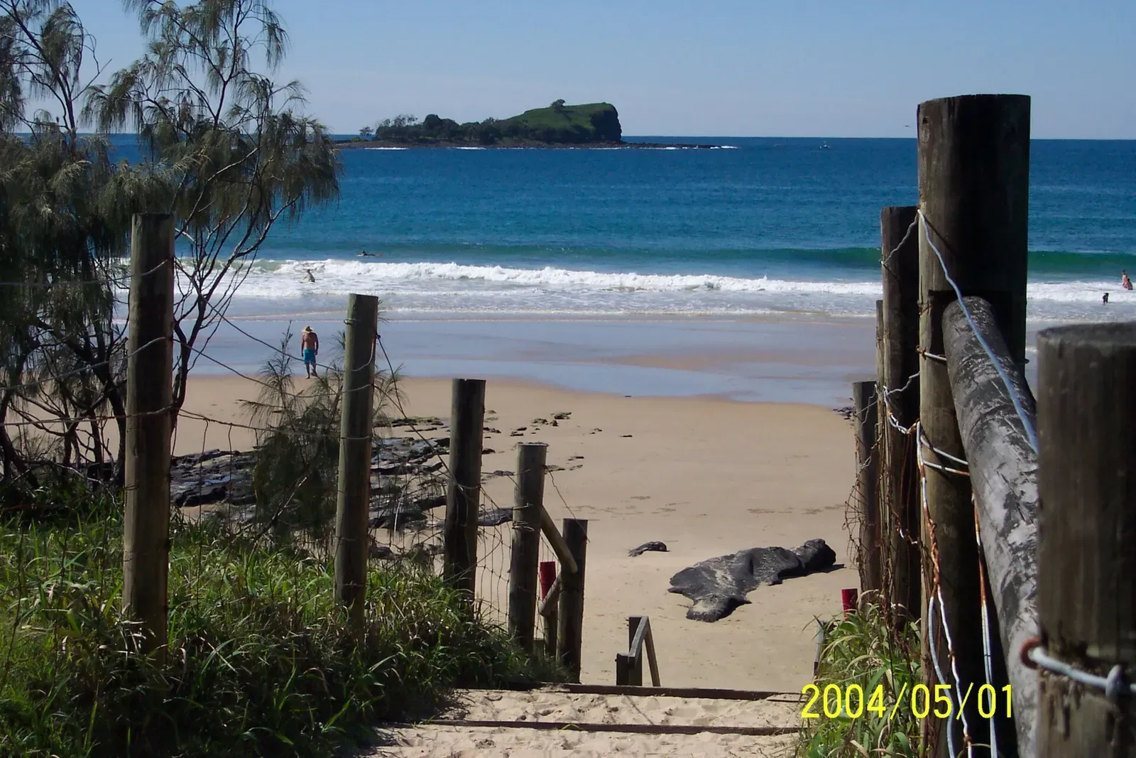 Daytime view of Mudjimba Island across the water from Mudjimba Beach, with a wooden beach-access fence in the foreground.