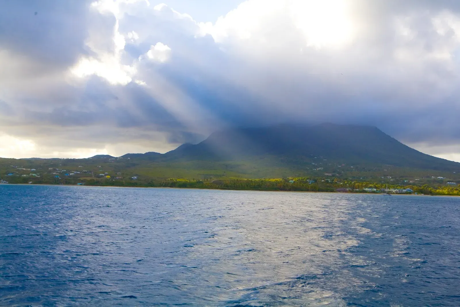 Sunbeams through heavy cloud over a green volcanic peak and a low coastline seen across dark blue sea — Nevis