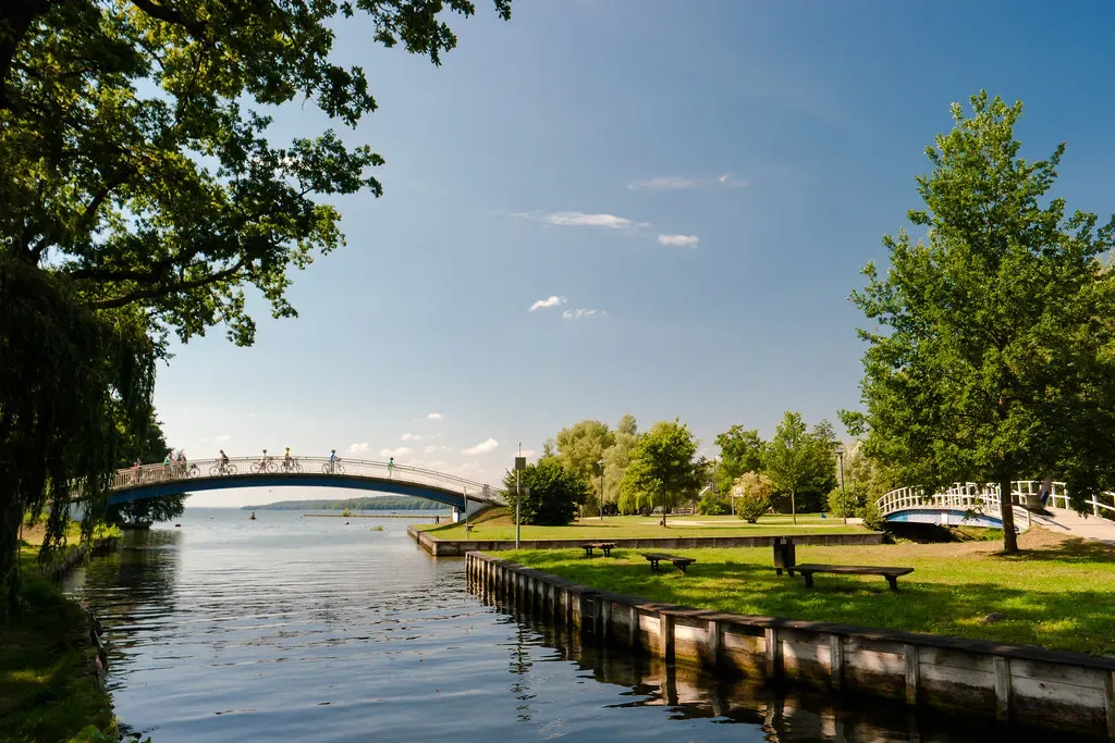 A calm channel lined with timber edging flows under an arched footbridge with cyclists toward open lake water, lawns and trees on both banks — Tollensesee & Neubrandenburg.