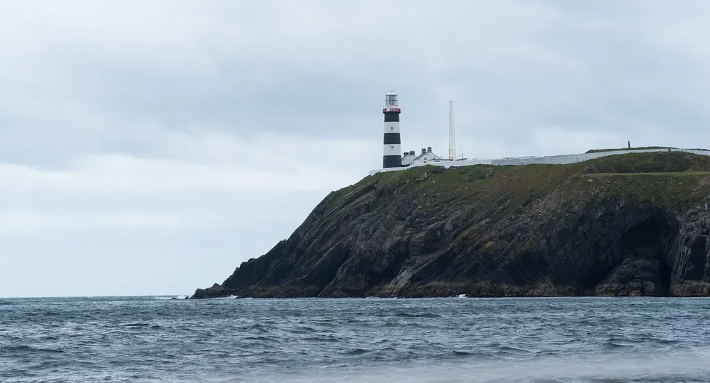 A black-and-white striped lighthouse stands on a dark green headland above a grey sea under an overcast sky — Kinsale & Old Head.