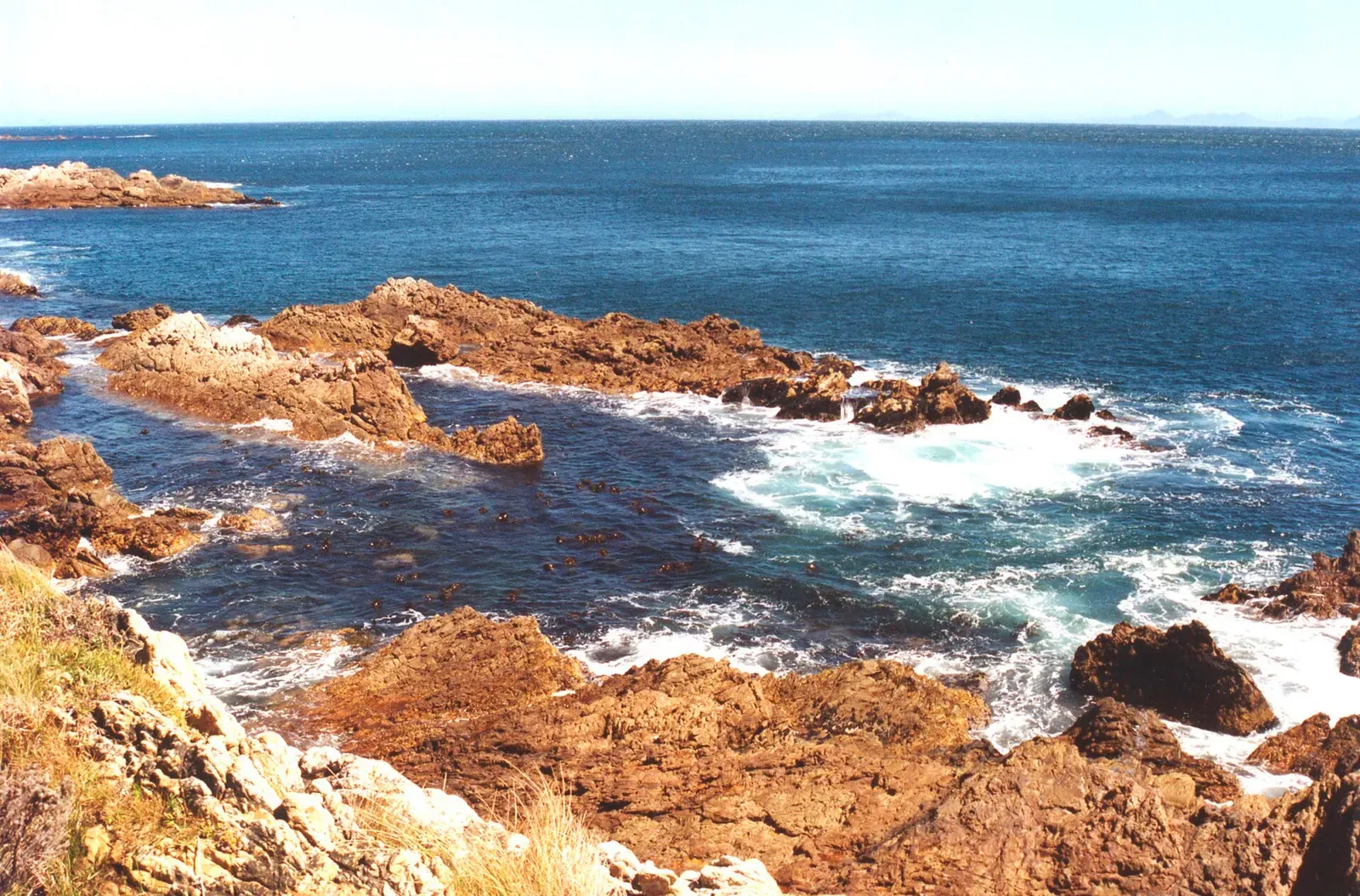 An orange rocky shoreline with a kelp-filled gully opening to a dark blue sea — Percy's Hole, Gordon's Bay & Rooi-Els