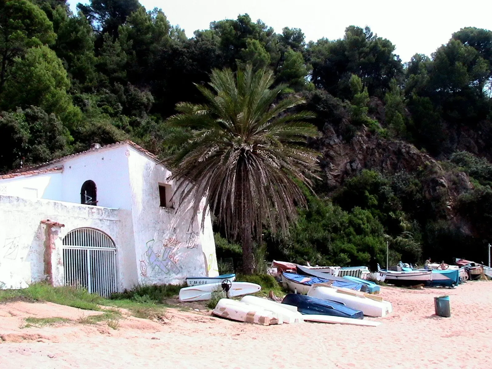 Whitewashed chapel and palm tree above small fishing boats hauled up on the sand at Cala Canyelles.