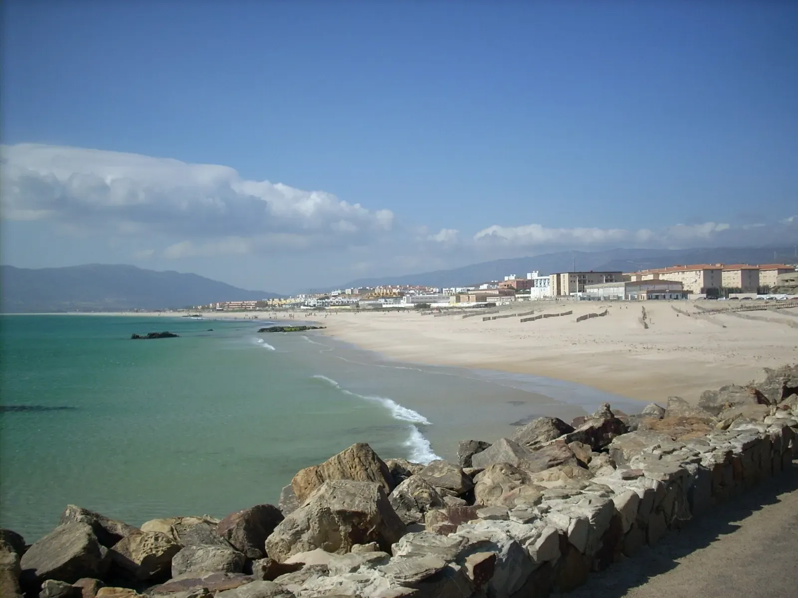 Rocky breakwater above a turquoise beach curving toward Tarifa's town and hills.