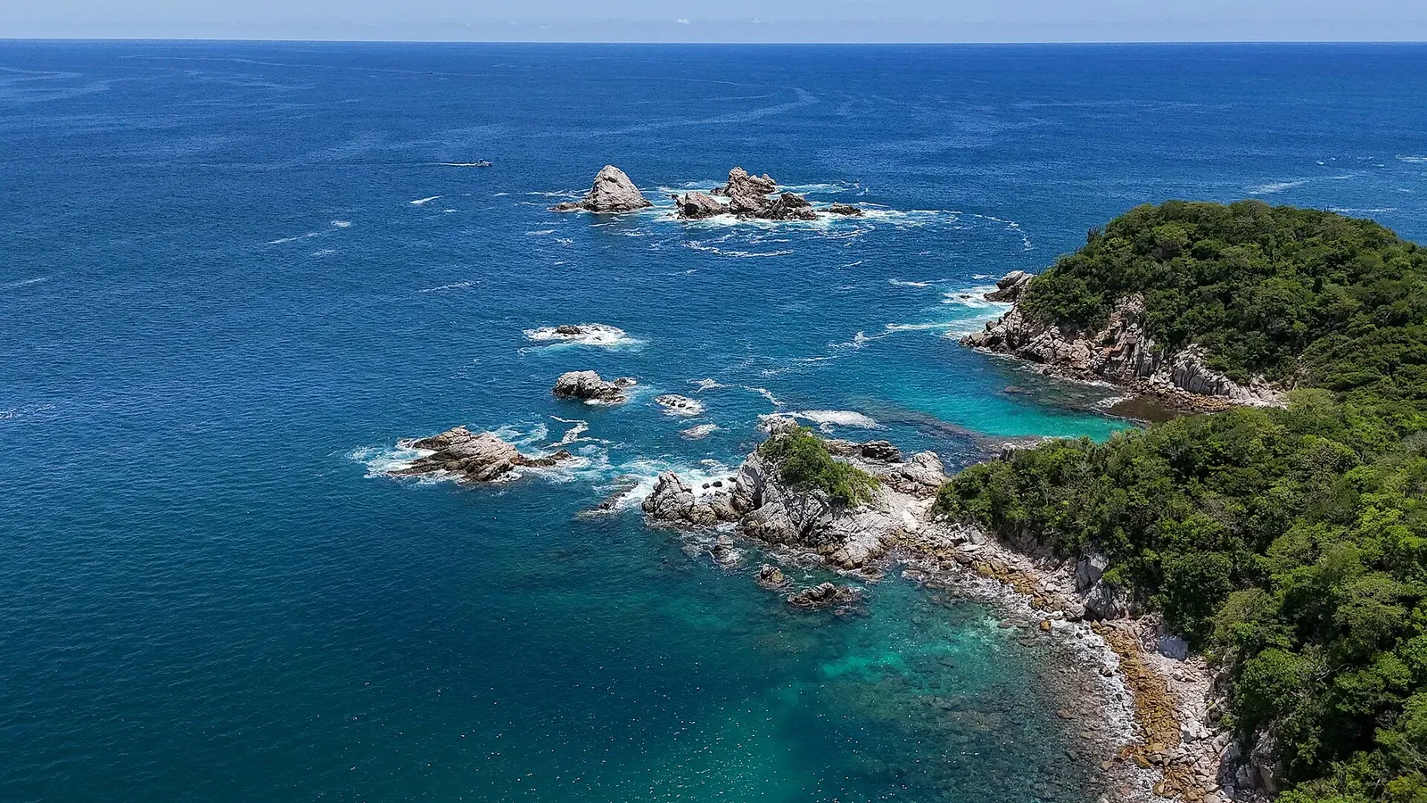 Aerial view of rocky islets and granite outcrops in clear blue water off a forested shore — Huatulco
