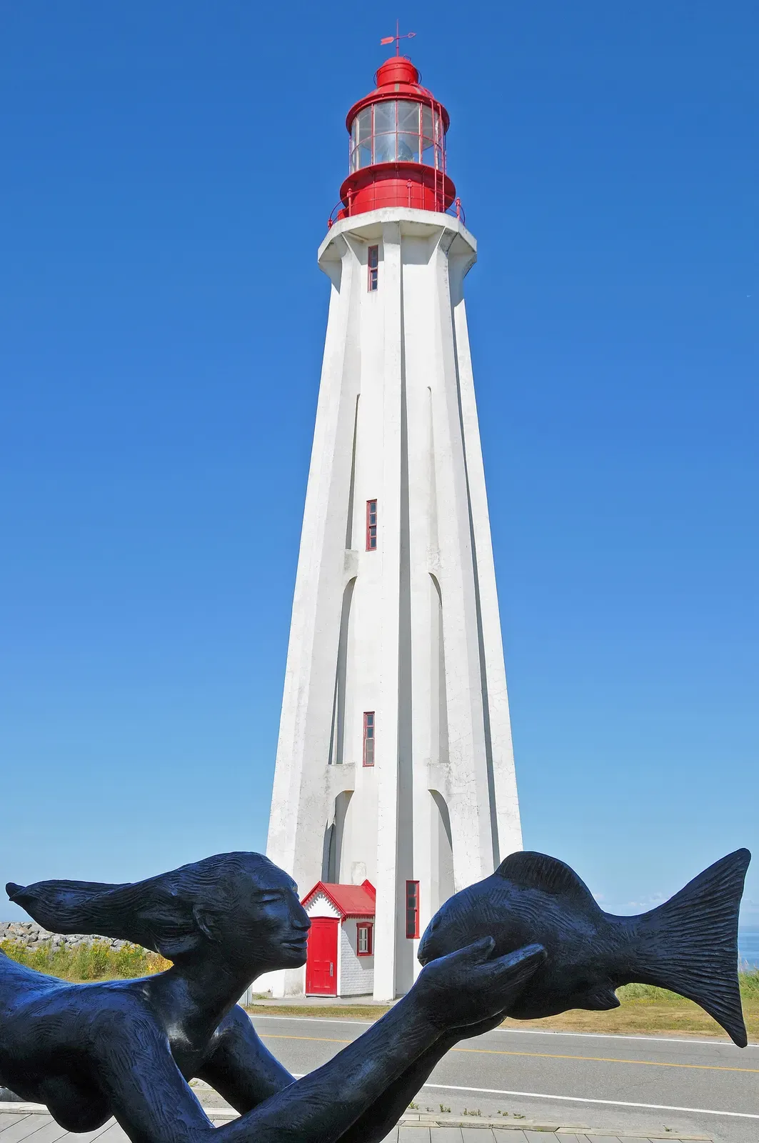 Base of a white lighthouse with two dark sculpted figures in front against a blue sky — Empress of Ireland, Rimouski