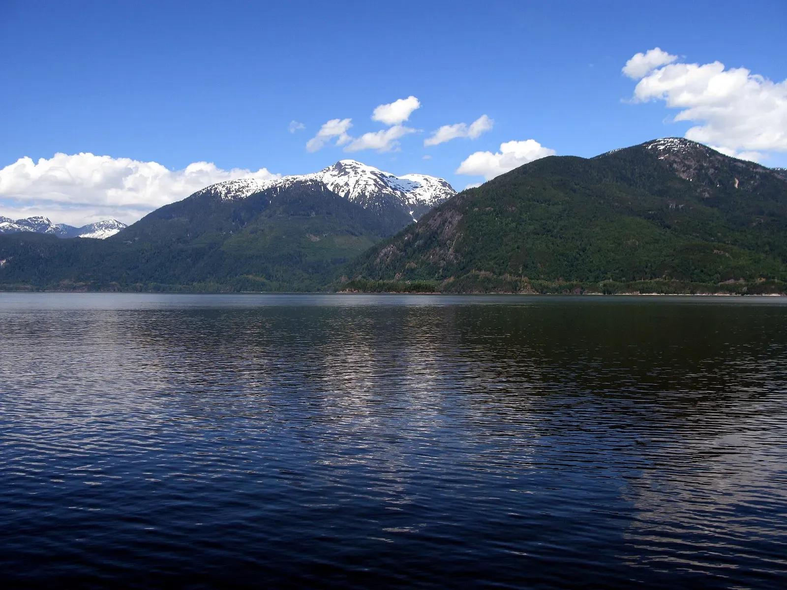 Calm blue water below forested mountains with snow-capped peaks under a blue sky — Porteau Cove, Horseshoe Bay & Howe Sound