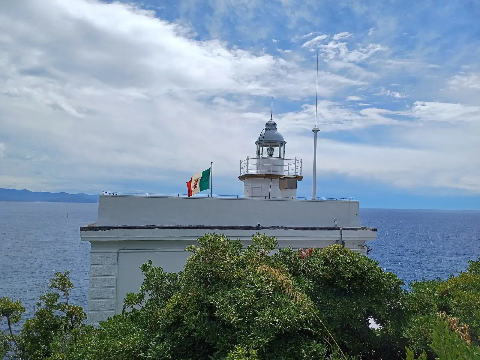 Whitewashed lighthouse building with an Italian flag, seen from below among trees — Punta del Faro, Portofino