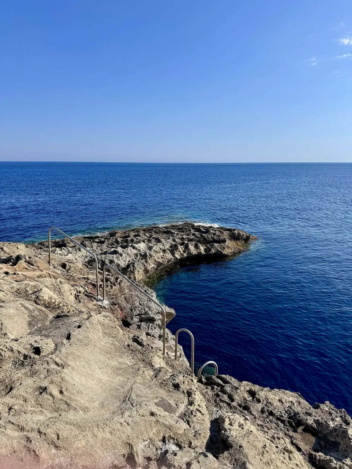 A rocky limestone headland with a metal ladder runs out into deep blue sea under a clear sky — Reqqa Point, Gozo & Malta.