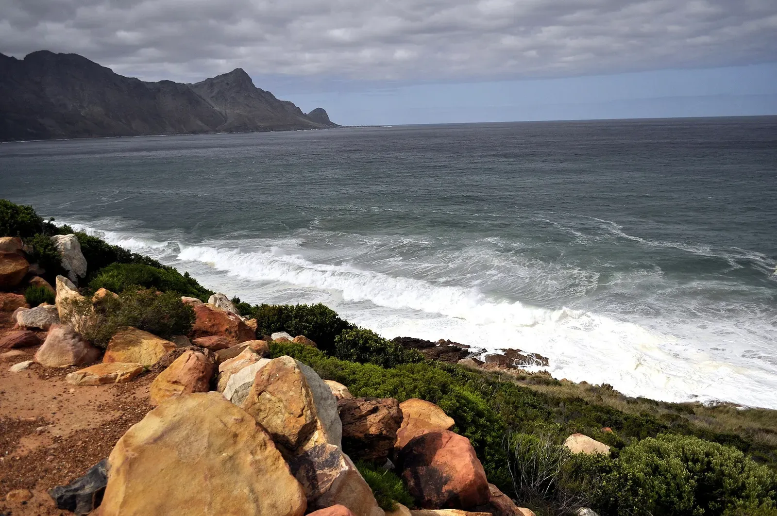 Rough grey sea and breaking surf below orange boulders, with a headland on the horizon — Rooi-els Point, Gordon's Bay & Rooi-Els
