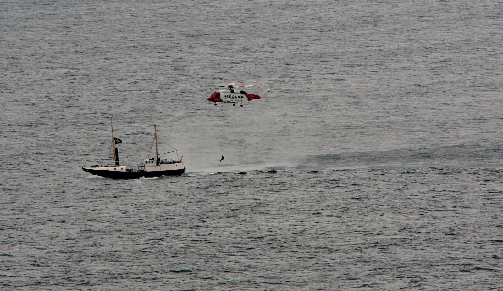 A red and white helicopter hovers low over grey sea beside a small two-masted boat — Shetland