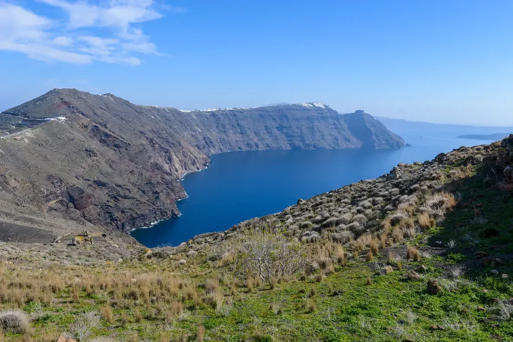 Steep layered brown cliffs dropping into calm deep blue water, seen from a grassy hillside under a clear sky — Santorini & the Caldera