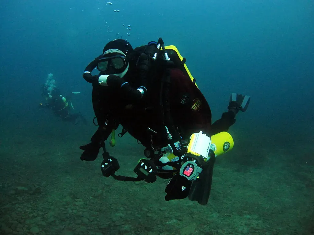 A rebreather diver in black faces the camera above a grey stony quarry floor, with a second diver in the green haze behind — Capernwray (Jackdaw Quarry), Capernwray & the Lake District