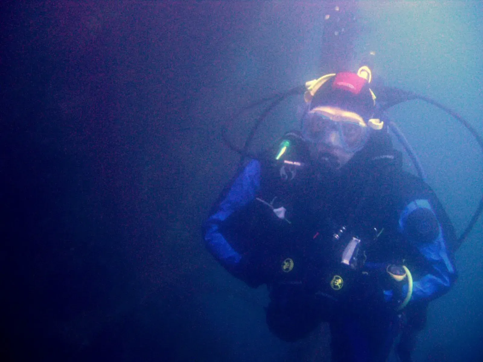 A diver in a black drysuit and a mask, lit by a torch, swims through deep blue water — James Eagan Layne, Plymouth Sound & HMS Scylla