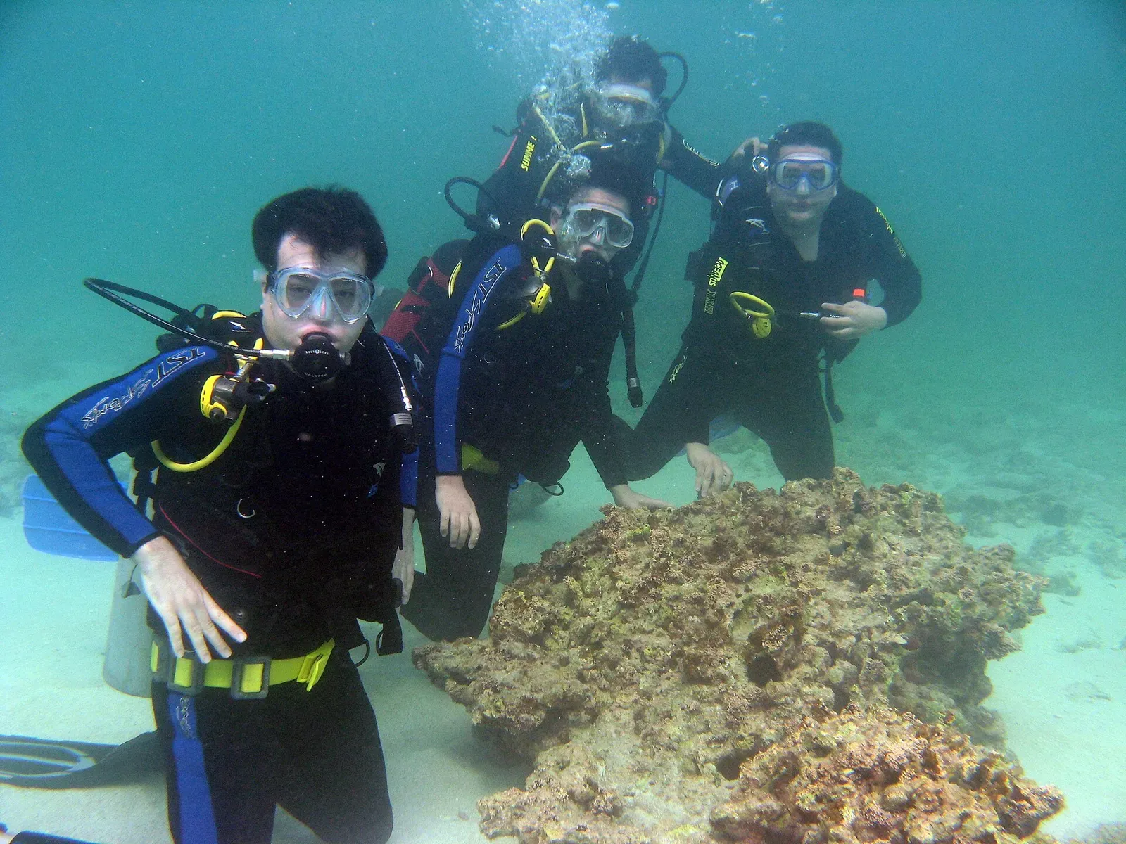 Four scuba divers in wetsuits and masks kneel on pale sand beside a brown coral head in green-blue water — Kish Island