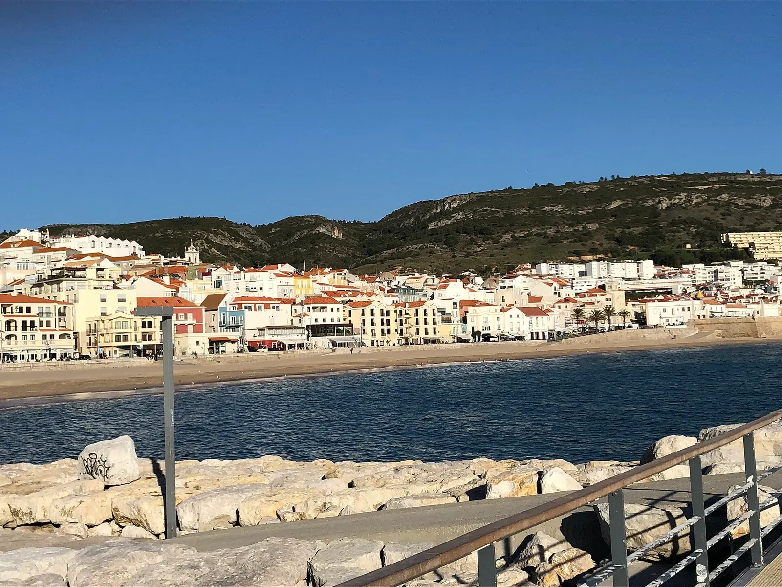 Red-roofed buildings along Sesimbra's beachfront, backed by wooded hills under a clear sky.