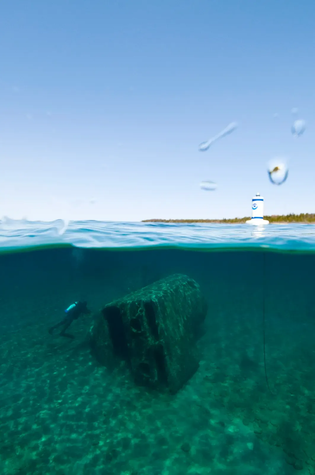 Split-level shot of a diver by a channel marker over the shallow wreck, water surface visible above — Monohansett, Thunder Bay