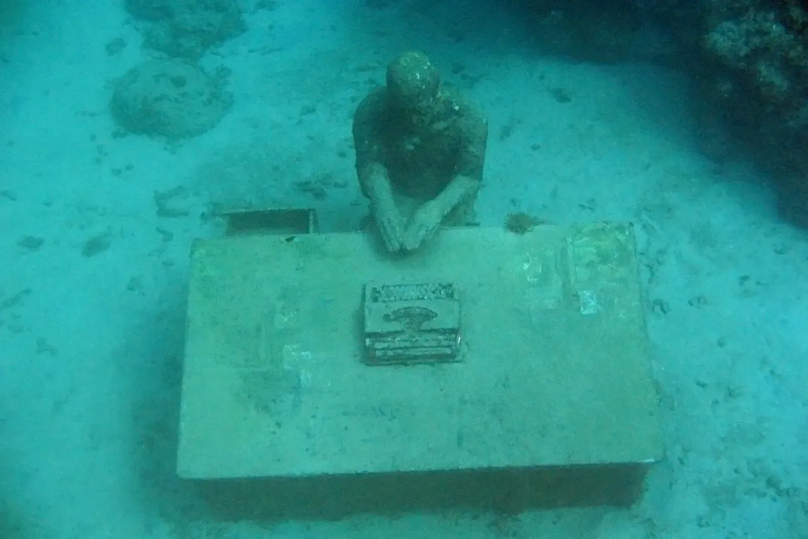 A cast figure seated at a typewriter desk on the seabed, sponges growing on its shoulders — Underwater Sculpture Park, Grenada
