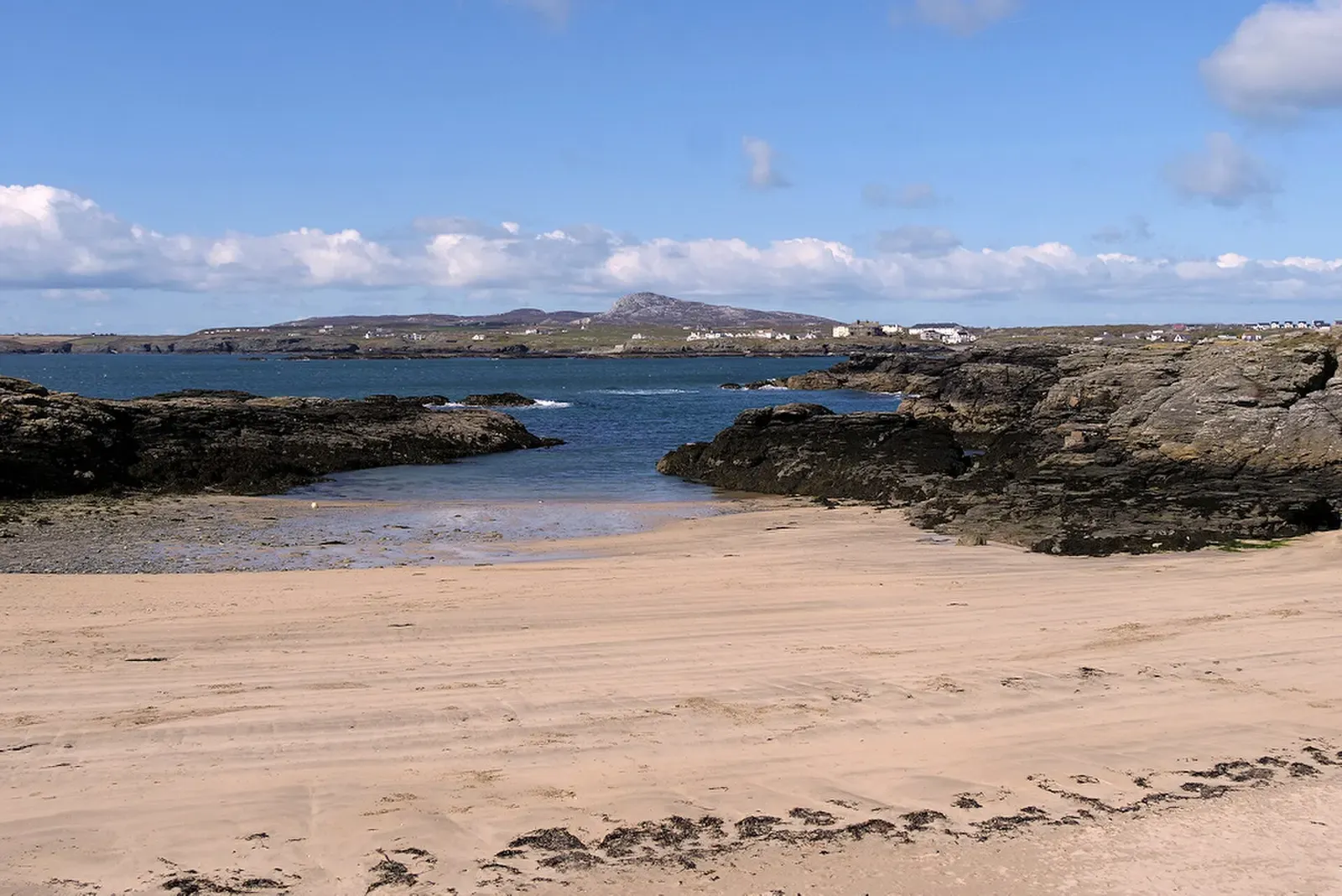 A small sandy cove with dark rocks at the water's edge and a hill on the far shore — Porth Castell, Anglesey