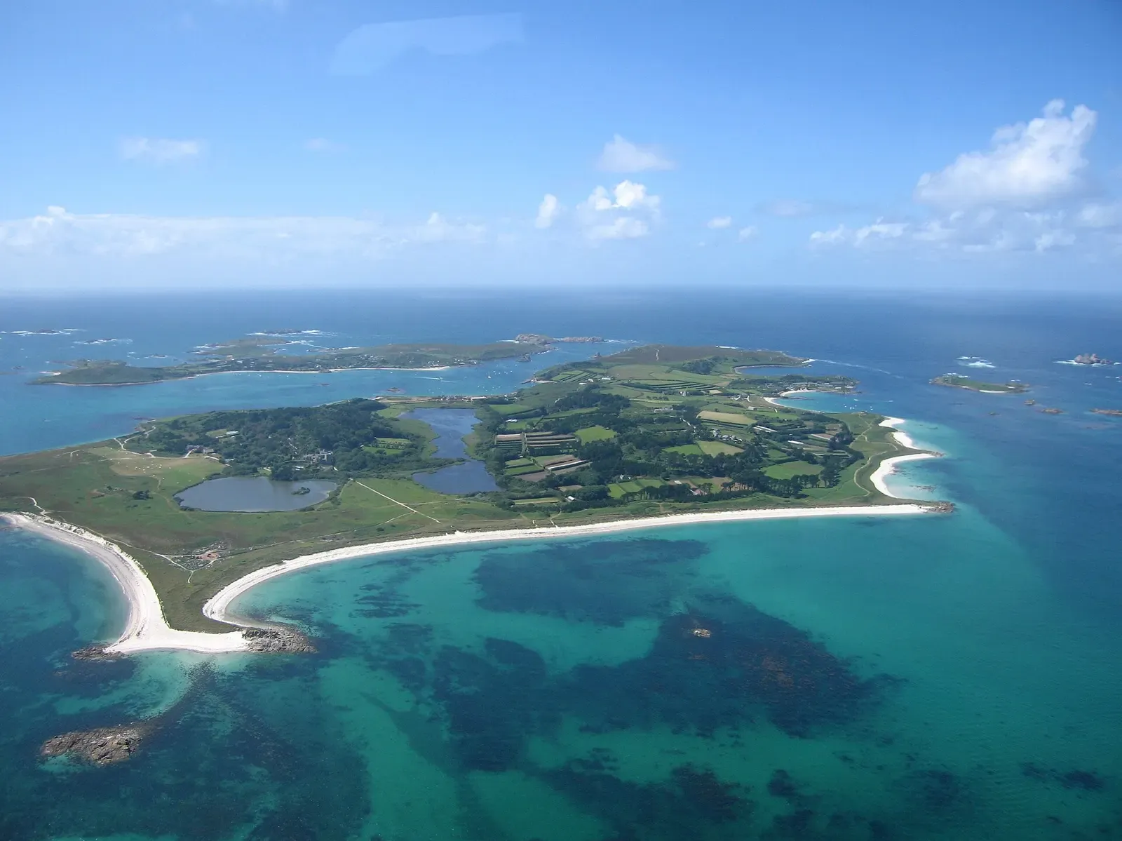 Aerial view of a green island with white sand beaches and turquoise shallows, more islands scattered across the sea behind — Isles of Scilly