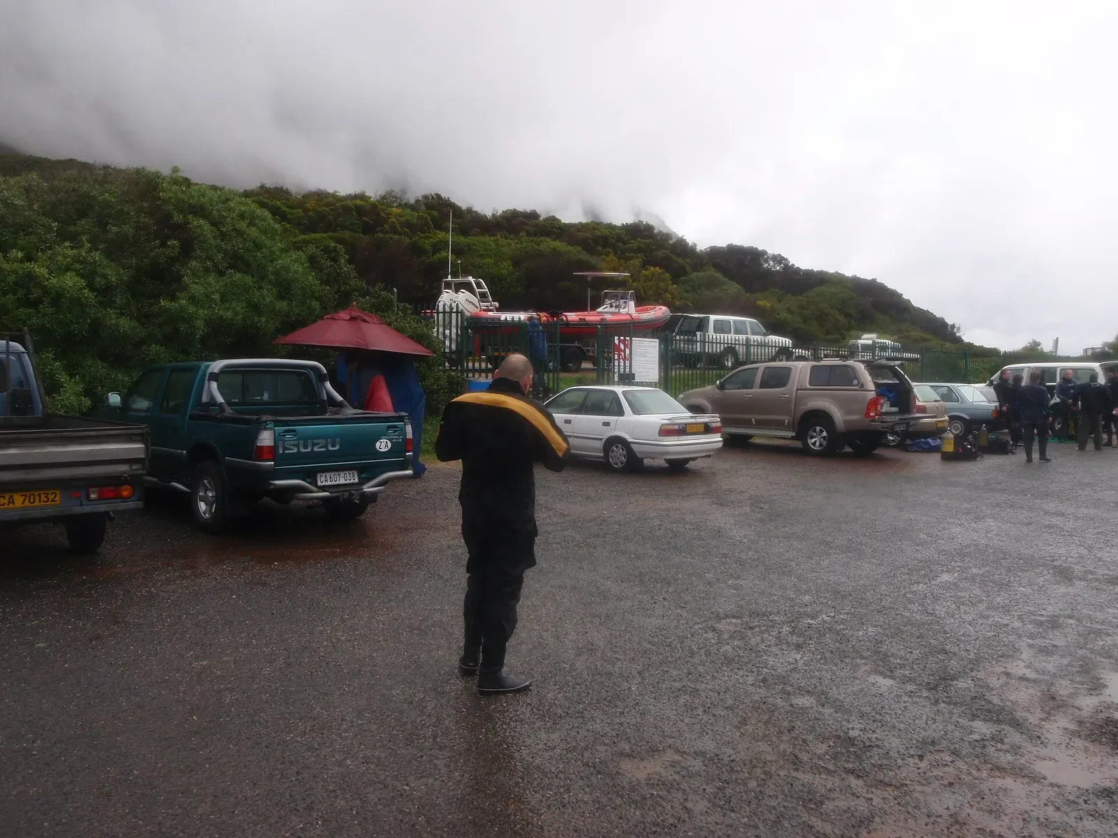 A diver in full kit walks across the slipway parking lot at Miller's Point under low cloud — Cape Town
