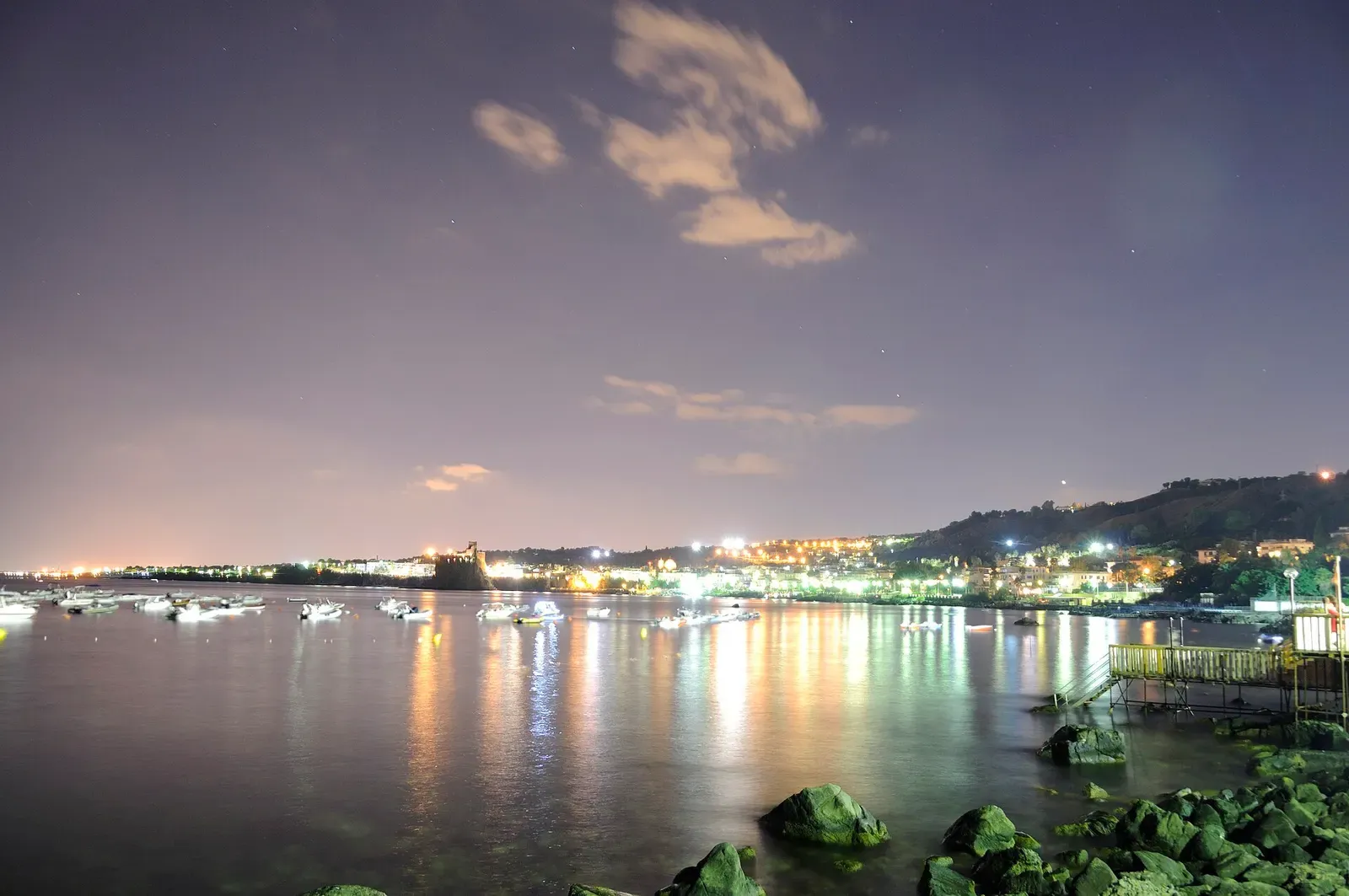 Boats sit in calm water at dusk in the harbour at Acitrezza, with the town's lights reflected on the surface.