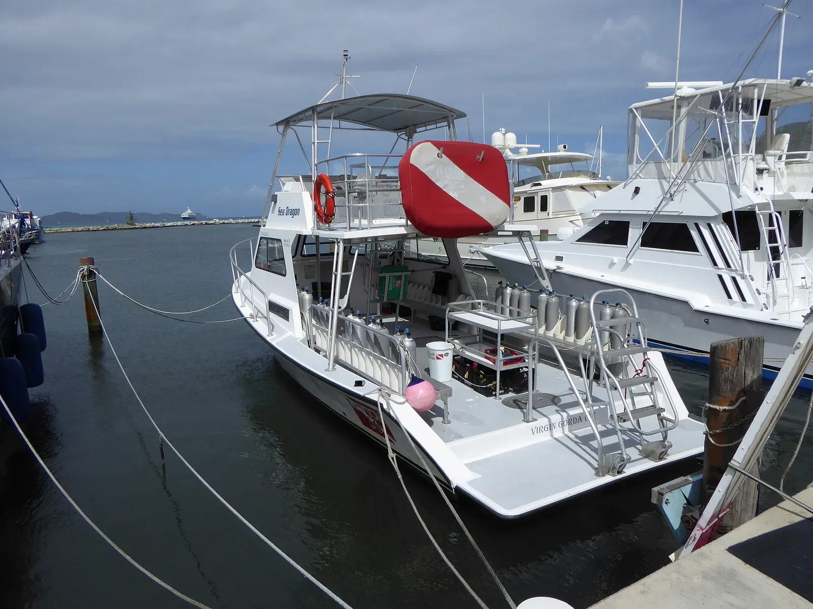 A white dive boat with racked scuba tanks, a boarding ladder and a large red-and-white dive flag, moored at a marina pier — Virgin Gorda & the Dog Islands.