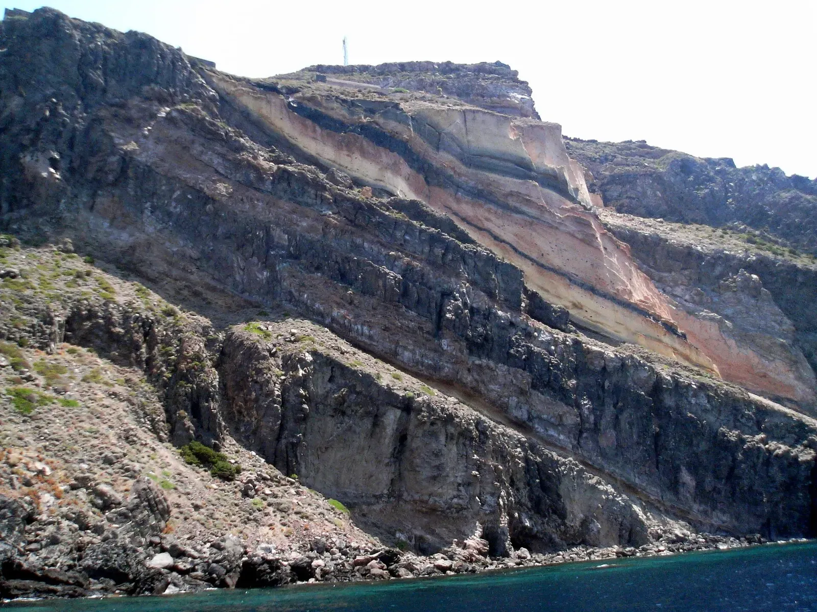 Steep layered volcanic cliffs in black, grey, cream and pink bands dropping to dark blue sea — Pantelleria