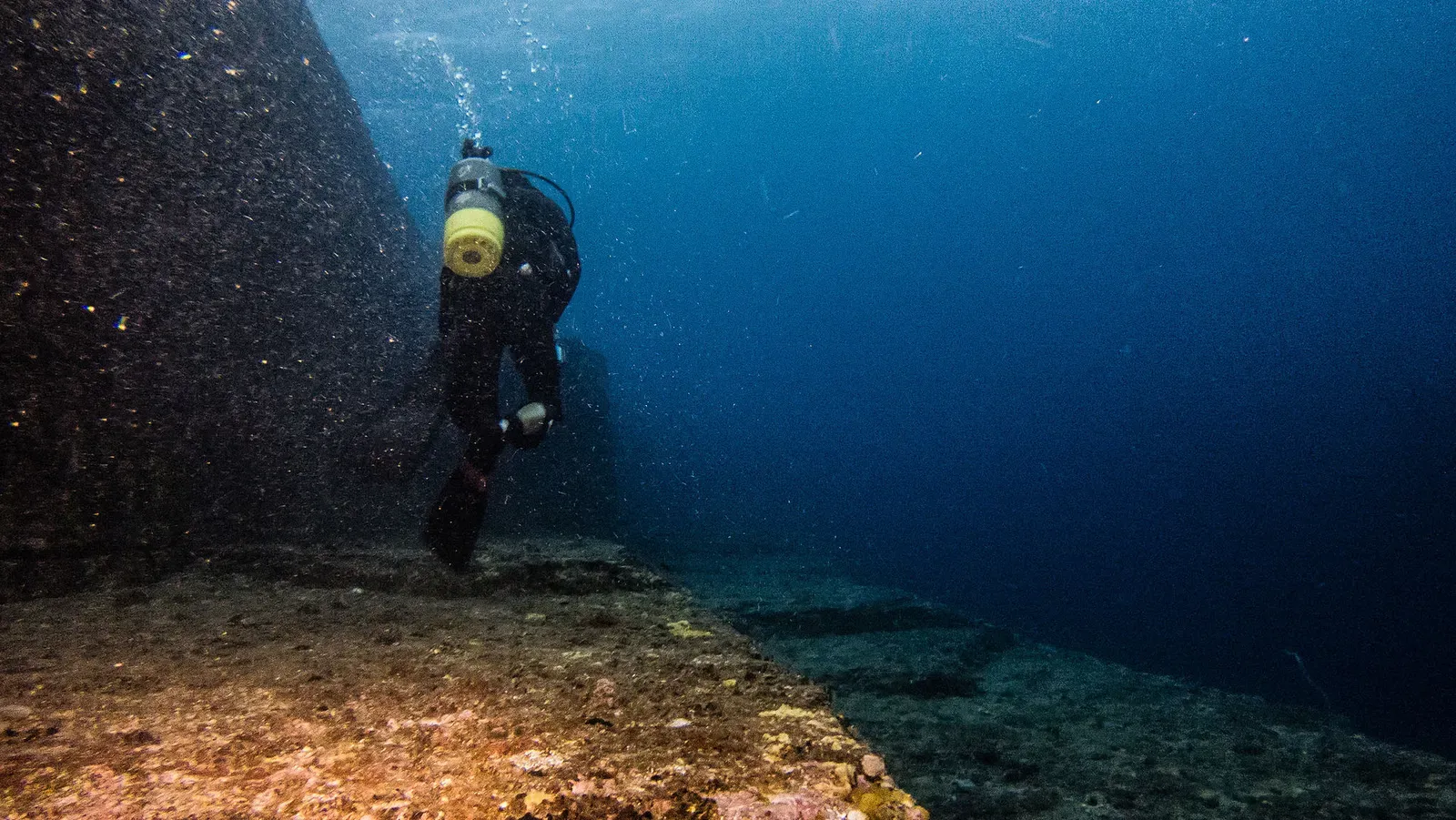 A diver swims along a sheer underwater rock wall at Yonaguni Monument, trailing bubbles.
