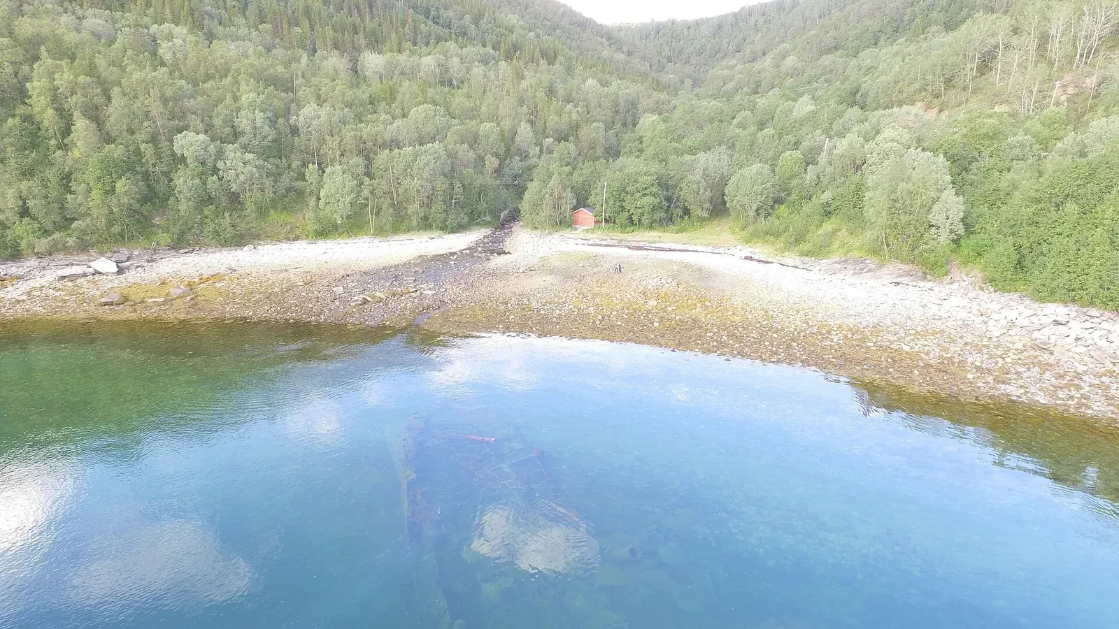 Aerial view of a rocky, tree-lined shoreline with the wreck faintly visible in the shallows at low tide — Hermann Künne, Narvik