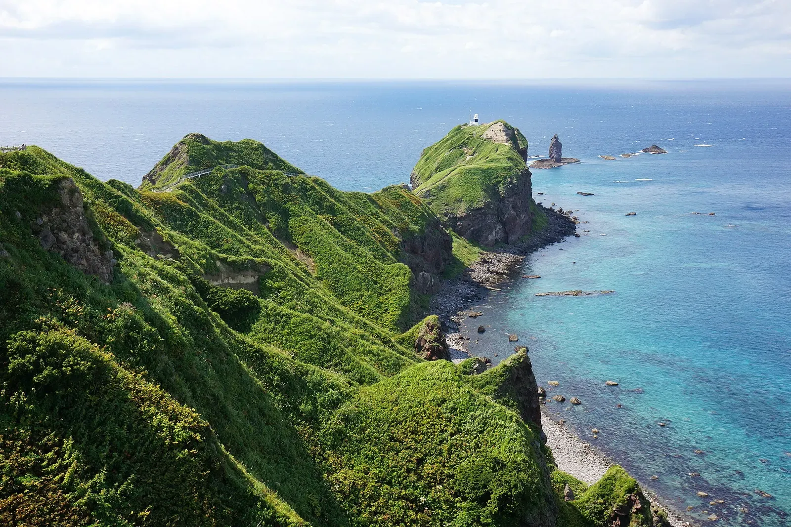 Green grassy ridges running out to a rocky cape with a sea stack, edged by clear turquoise shallows — Shakotan Peninsula