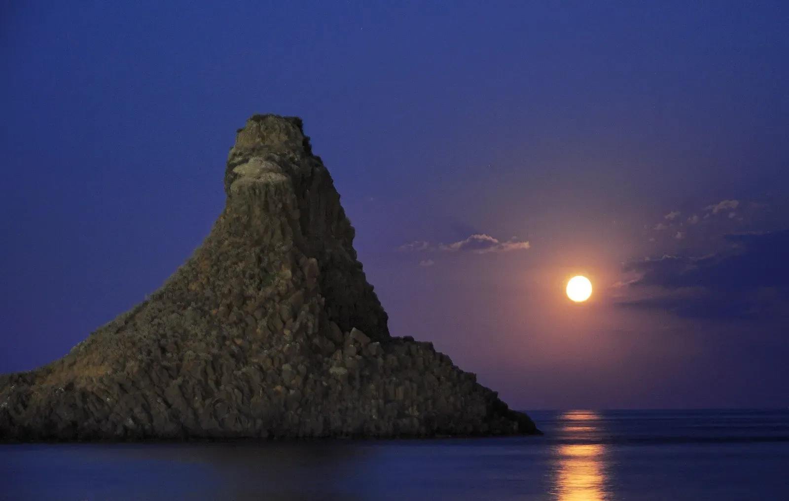 A pointed sea stack is silhouetted against the sky as the moon rises low over the water beside it.