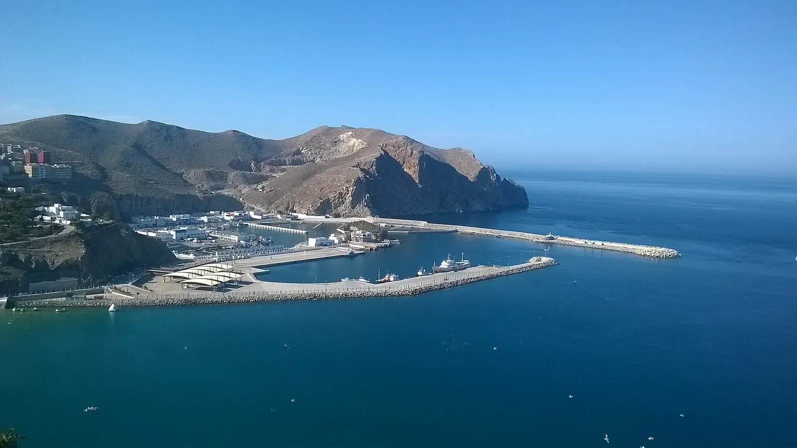 Harbour with long breakwaters and moored boats below a steep rocky headland and deep blue sea — Al Hoceima
