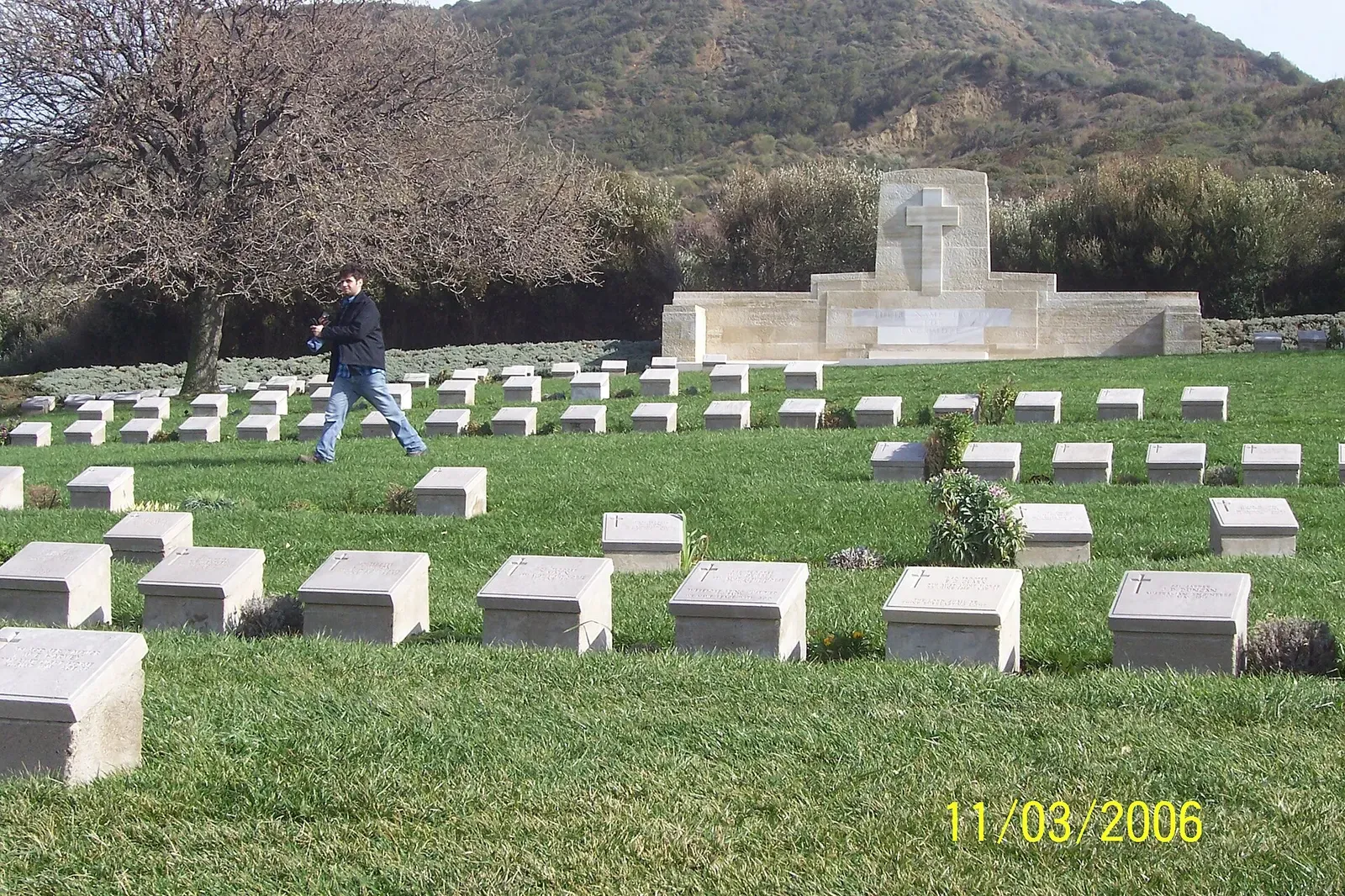 Rows of white headstones in a lawn cemetery at Anzac Cove with scrub-covered hills behind — Gallipoli.