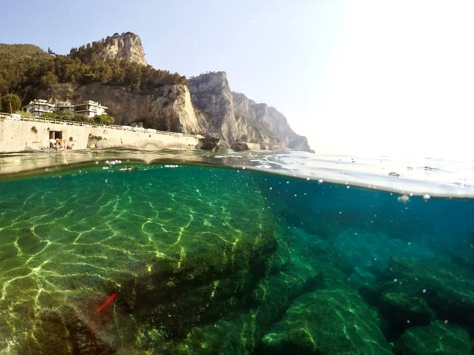 Over-under shot of submerged beachrock in clear turquoise water off Varigotti, with a limestone headland behind