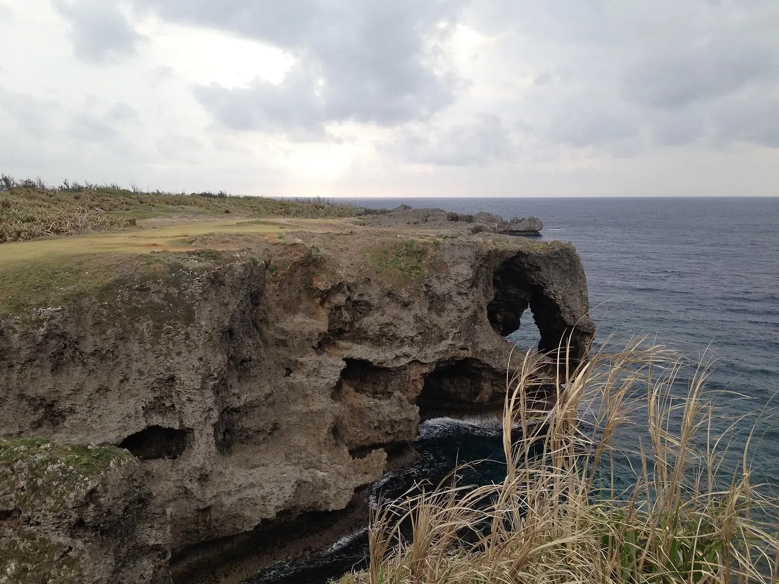 A wide view across the grassy clifftop of Cape Manzamo toward its coral-rock arch — Horseshoe Cliff, Okinawa.