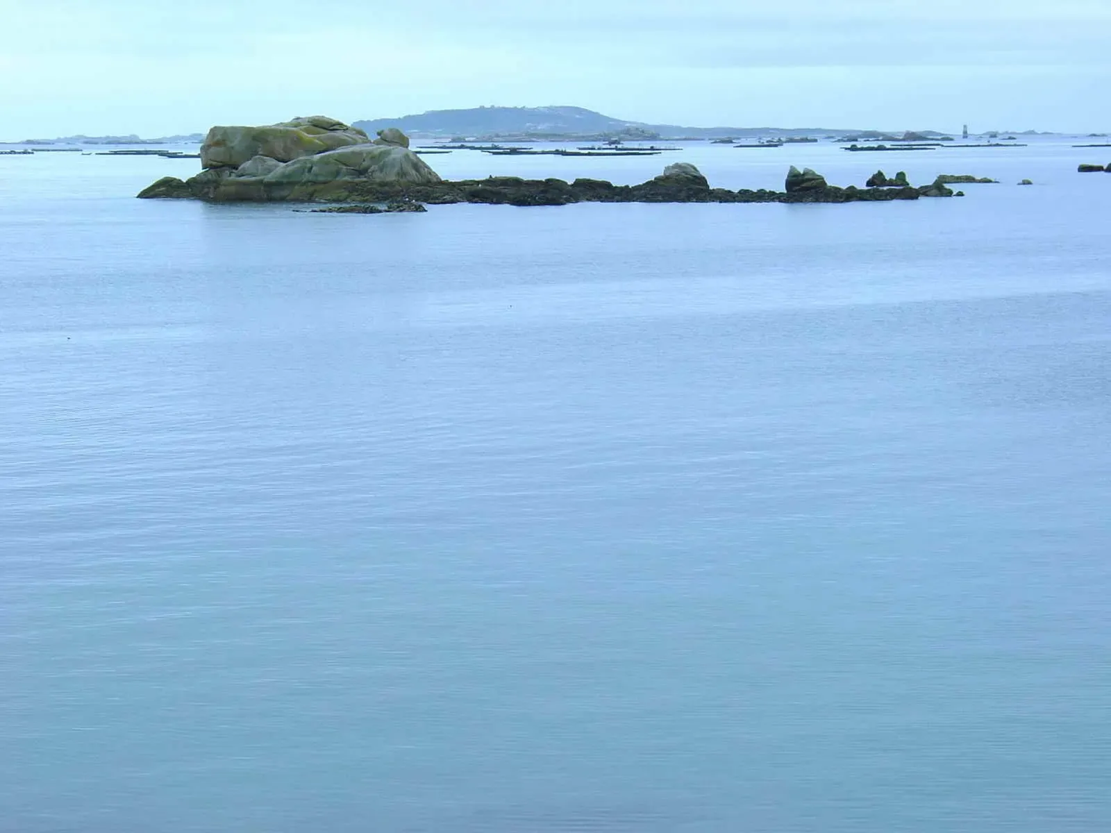 Granite boulders on a rocky islet in flat water, with a low island and floating rafts in the distance — Sálvora, Ons & Ría de Arousa