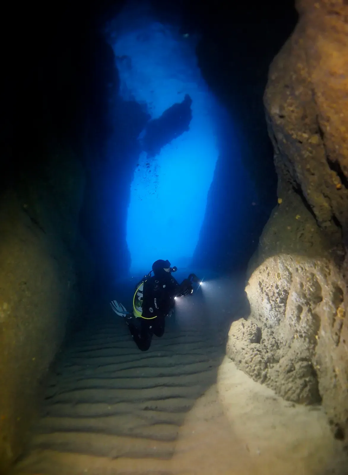 A diver with a torch swims through a narrow, blue-lit cavern with rippled sand on its floor — Mgarr ix-Xini, Gozo & Malta.