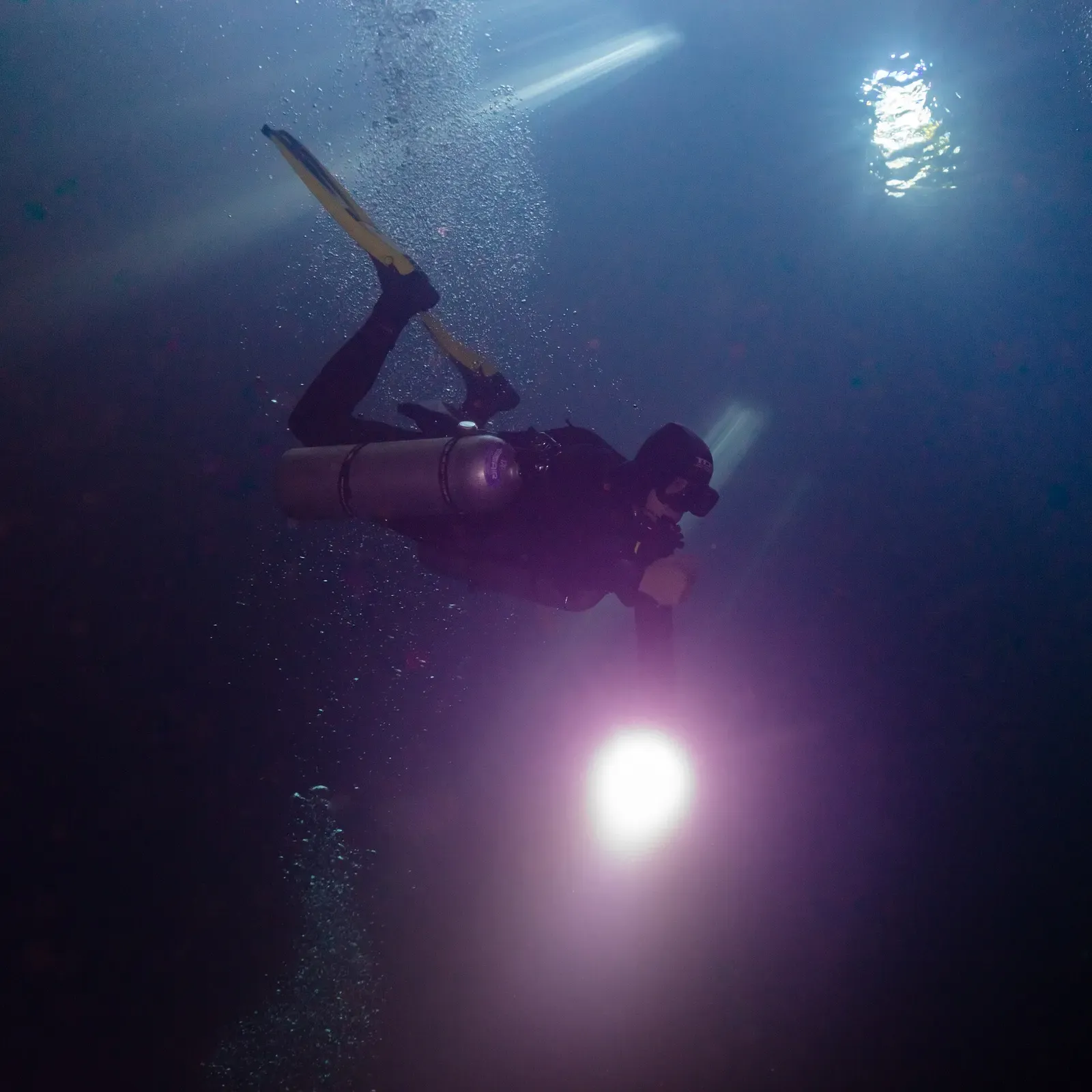 Diver with a bright torch in near-dark blue water deep in the shaft